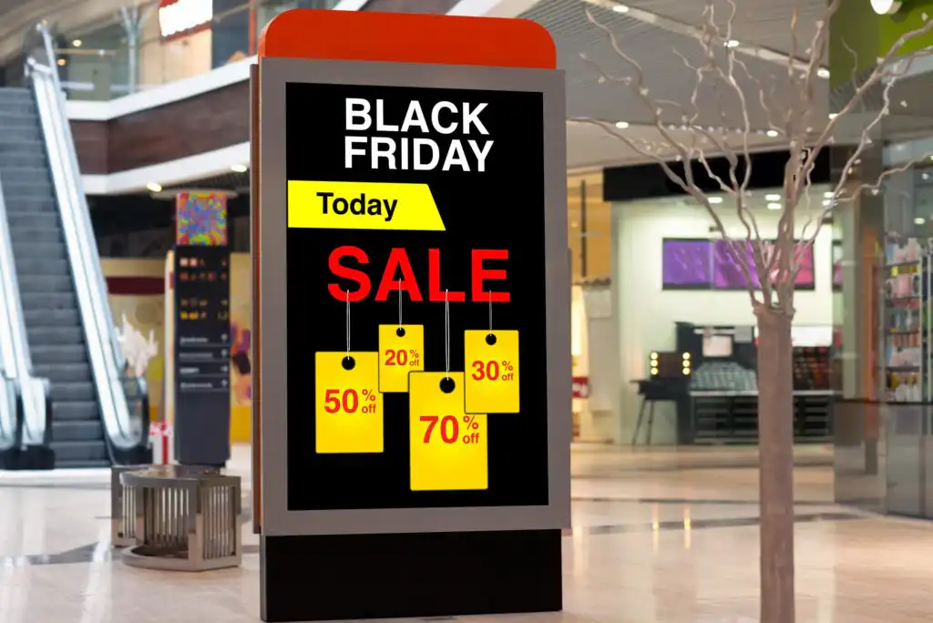 A digital sign in a shopping mall displays Black Friday Today SALE with discount tags showing 50%, 70%, 20%, and 30% off. An escalator and store fronts are visible in the background.