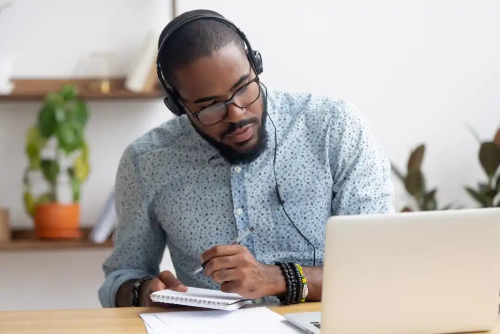 A man wearing headphones and glasses sits at a desk, looking at a laptop screen while taking notes in a notebook. There are plants and shelves in the background.