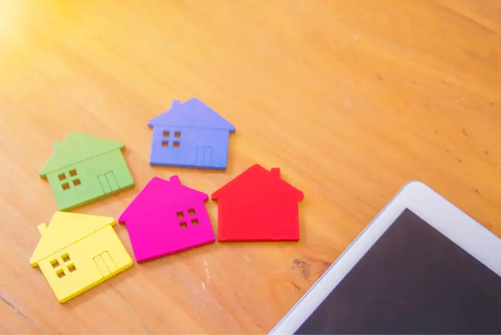 Colorful paper cutouts of five small houses (yellow, green, blue, pink, and red) are arranged on a wooden surface next to the corner of a digital tablet.