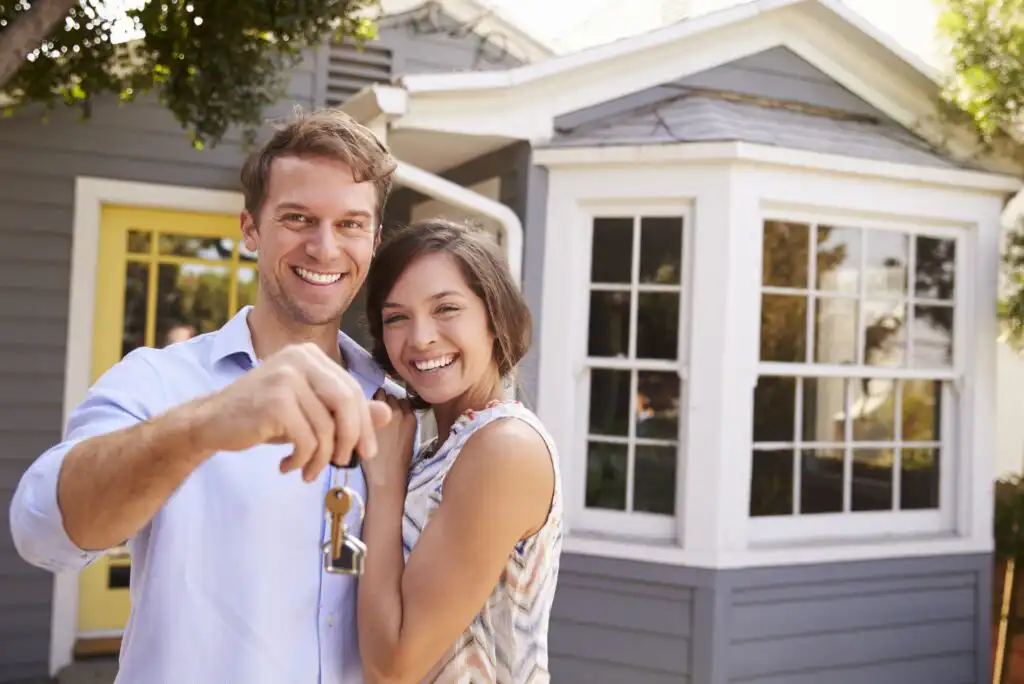 A smiling couple stands in front of a house, holding up a key, suggesting they are new homeowners. The house has gray siding, large windows, and a yellow door.