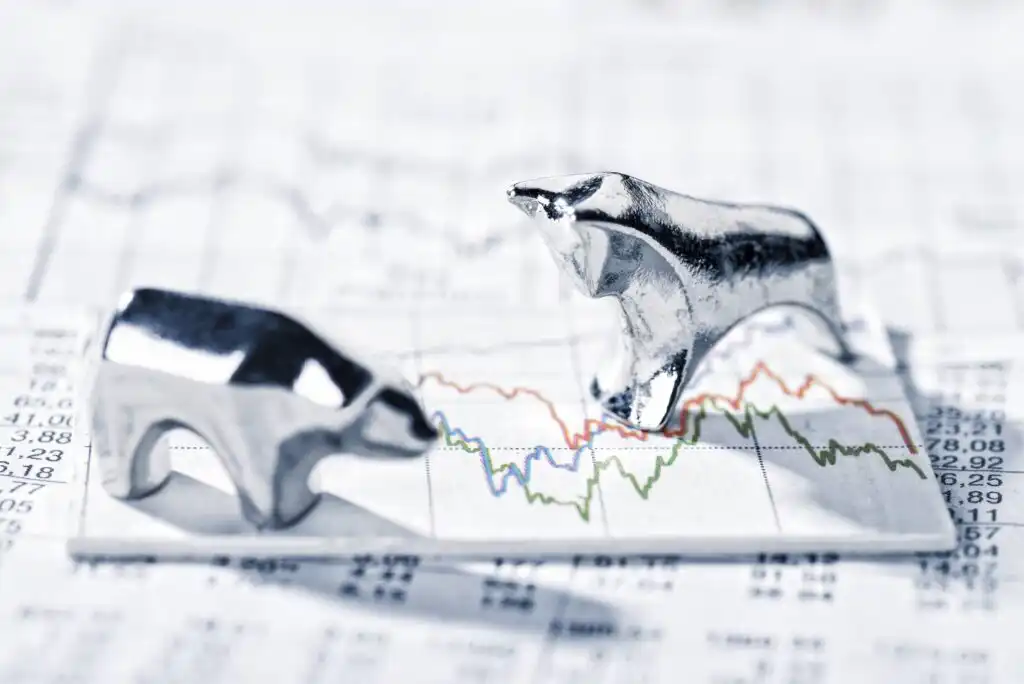 Two metallic figurines, a bull and a bear, face each other on top of a paper with financial graphs and stock market data, symbolizing bullish and bearish market trends.