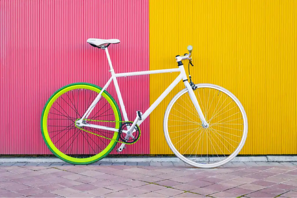 A white bicycle with a green rear wheel and white front wheel stands against a wall split into vertical pink and yellow halves. The ground is paved with light-colored tiles.