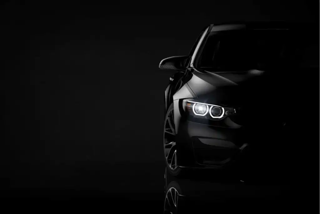 A sleek black car is shown in a dark setting with only the front half visible. Its headlights are on, highlighting the sharp contours and modern design of the vehicle against a black background.