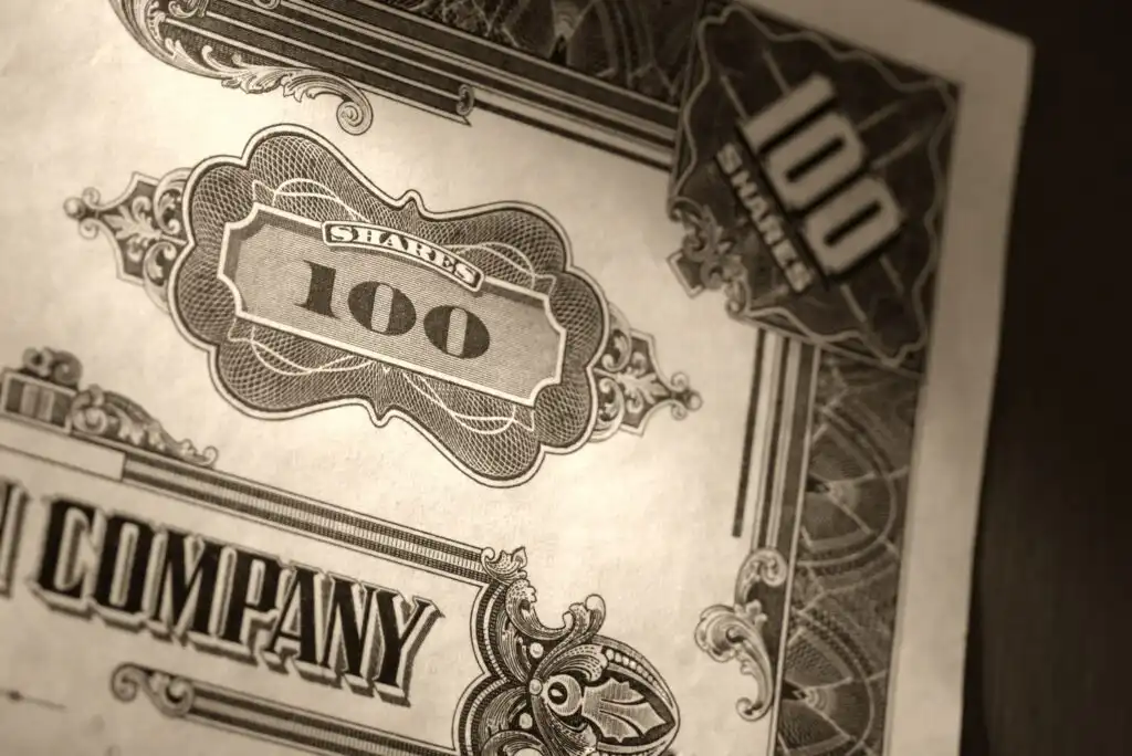 A close-up of an ornate, vintage-style stock certificate showing “100 shares” and decorative patterns, with the partially visible word “COMPANY” printed in bold letters.