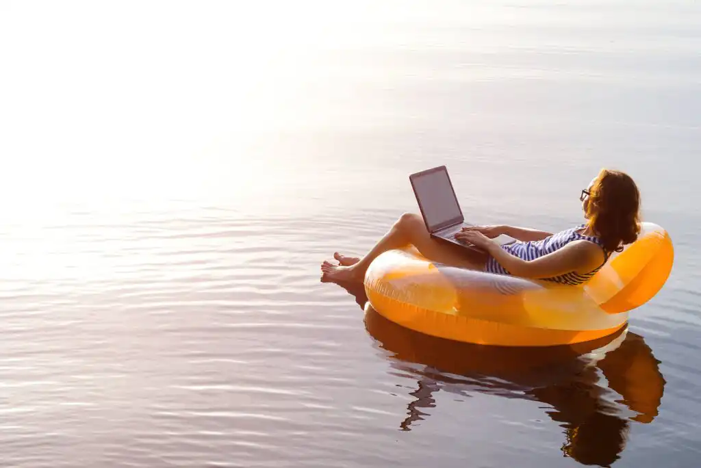 A person in sunglasses lounges on a yellow inflatable ring in calm water, using a laptop, with sunlight reflecting off the surface.