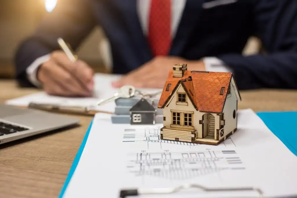 A miniature house, keys, and blueprints sit on a desk, with a person in a suit signing documents in the background, suggesting a real estate or mortgage transaction.