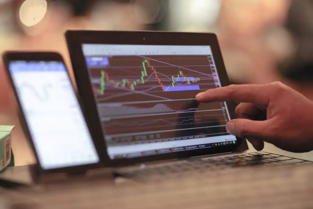 A person points to a candlestick chart with technical indicators displayed on a laptop screen, analyzing stock or cryptocurrency market data; a smartphone with charts sits nearby.