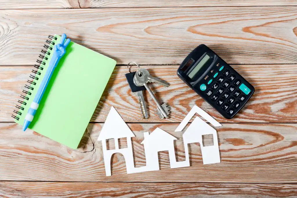 A green notepad with a blue pen, a set of keys, a black calculator, and paper cutouts of houses arranged on a wooden surface.