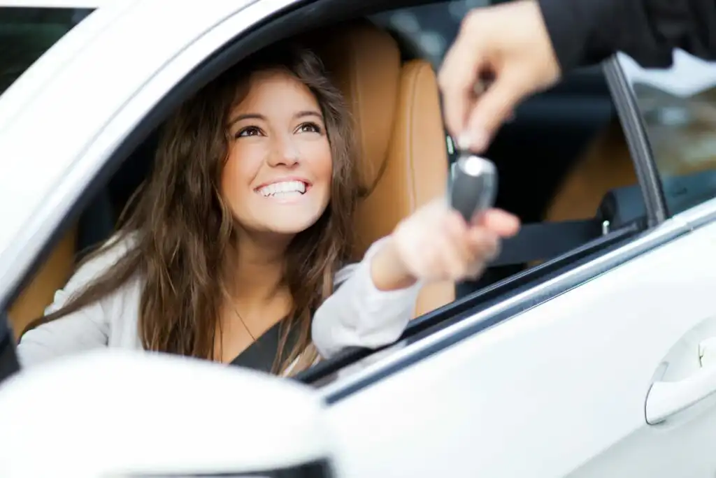 A smiling woman sitting in the driver’s seat of a car reaches out to receive a car key from someone standing outside the vehicle.