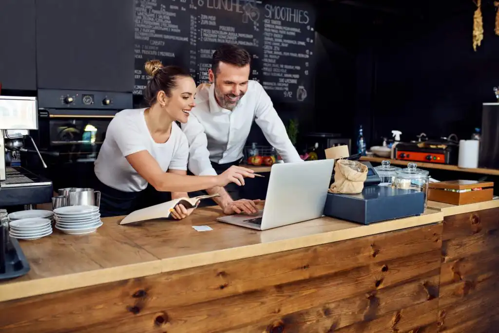 Two smiling people behind a café counter look at a laptop together. One is holding a notepad and pointing at the screen. Plates, cups, and bakery items are on the wooden counter, with a chalkboard menu in the background.