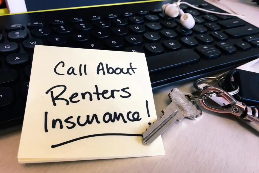 A sticky note reading Call About Renters Insurance! sits on a desk next to a computer keyboard, a pair of white earbuds, and a key on a keyring.