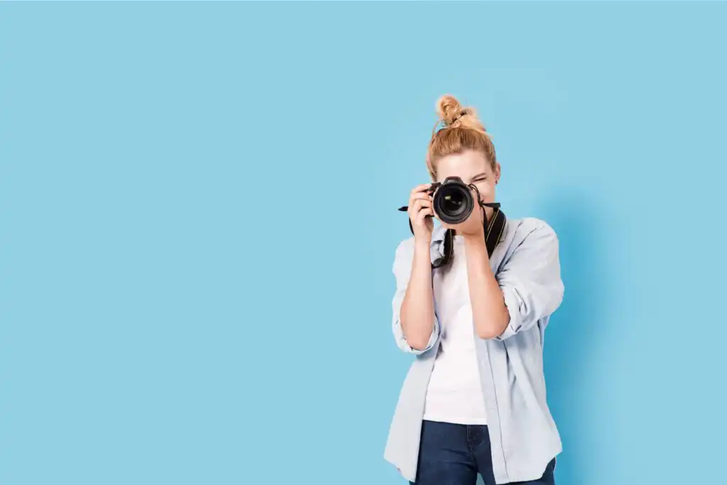 A person with blonde hair in a bun, wearing a light blue shirt and jeans, is standing against a light blue background and holding a camera up to their face as if taking a photo.
