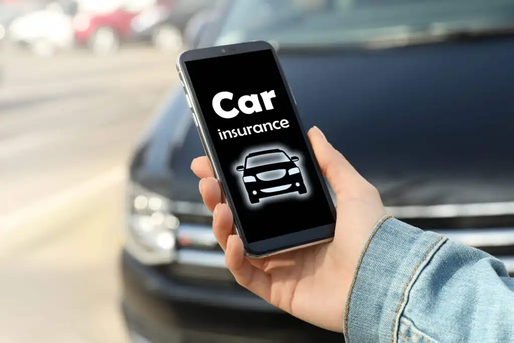 A person holds a smartphone displaying a screen with the words Car insurance and a car icon. A blurred black car and street are visible in the background.