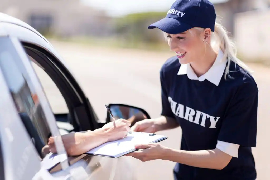 A smiling woman wearing a CHARITY t-shirt and cap holds a clipboard while a person inside a car signs a document. The scene is outdoors and appears to be part of a donation or volunteer event.