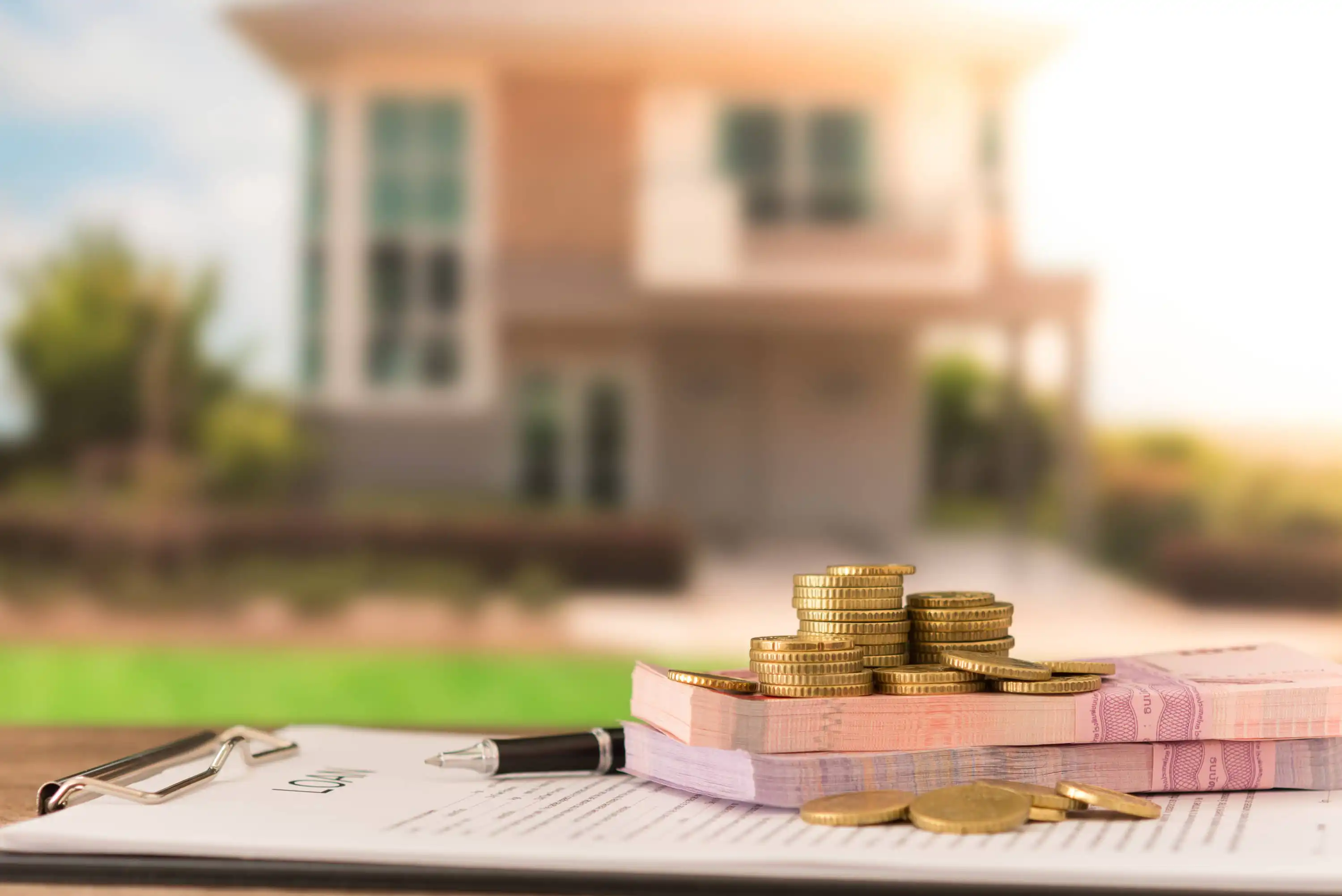A stack of coins and a bundle of banknotes sit on top of a clipboard with papers and a pen, with a blurred house in the background.