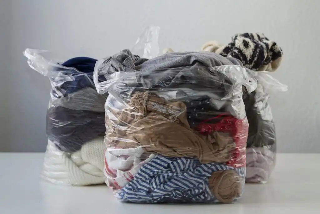Clear plastic bags filled with various folded clothes, including patterned, striped, and solid-colored garments, stacked on a white surface against a plain light background.
