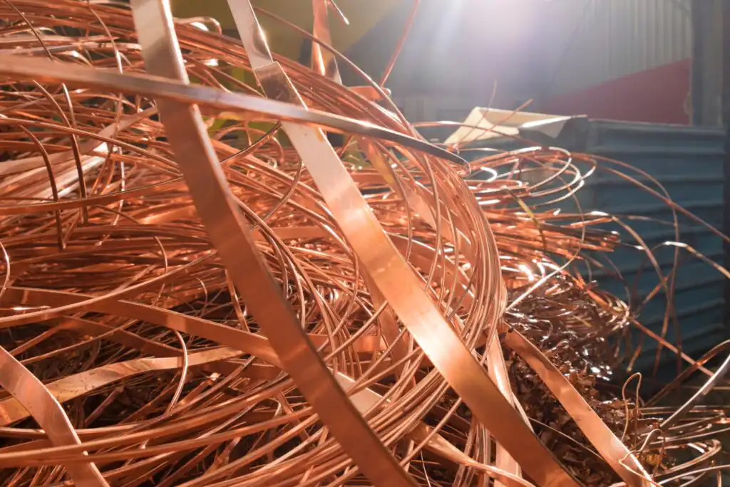 A close-up of a tangled pile of shiny copper wires and strips, illuminated by sunlight in what appears to be an industrial or workshop setting.
