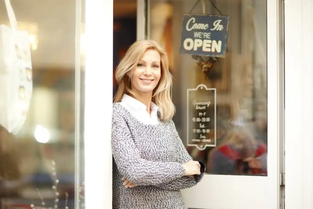 A woman with blonde hair, wearing a gray sweater and white shirt, stands smiling with arms crossed in front of a glass door with an “OPEN” sign and business hours visible.