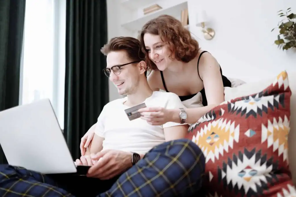 A smiling couple sits on a couch with a colorful patterned pillow. The man uses a laptop while the woman, leaning over his shoulder, holds a credit card as they shop online together.