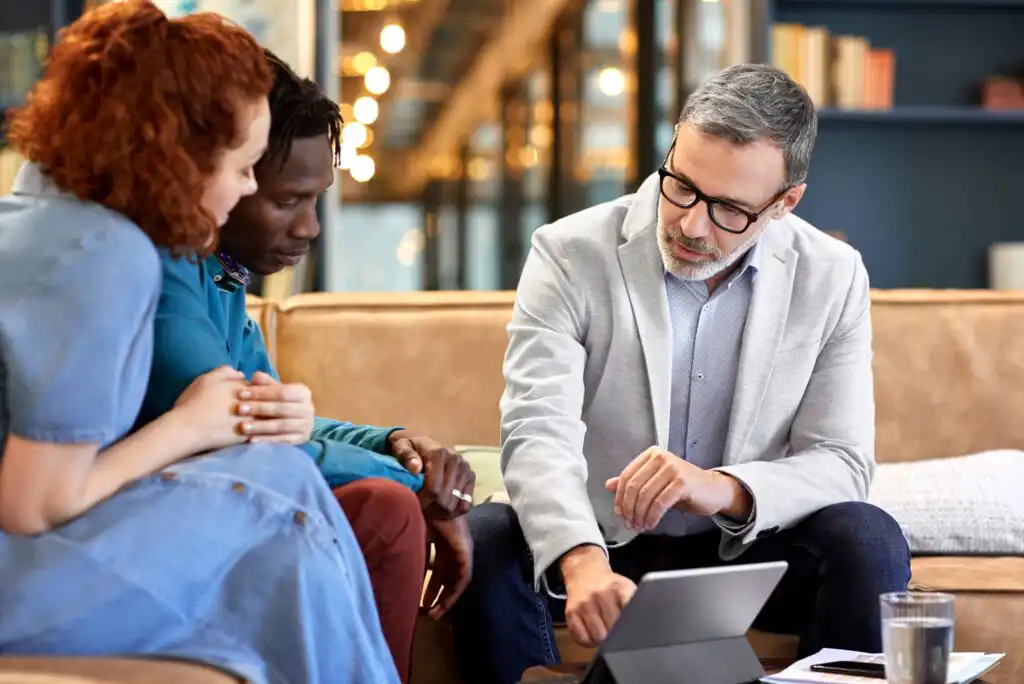 A man in glasses and a light blazer shows something on a tablet to a woman and a man sitting beside him on a couch in a modern office or lounge setting.
