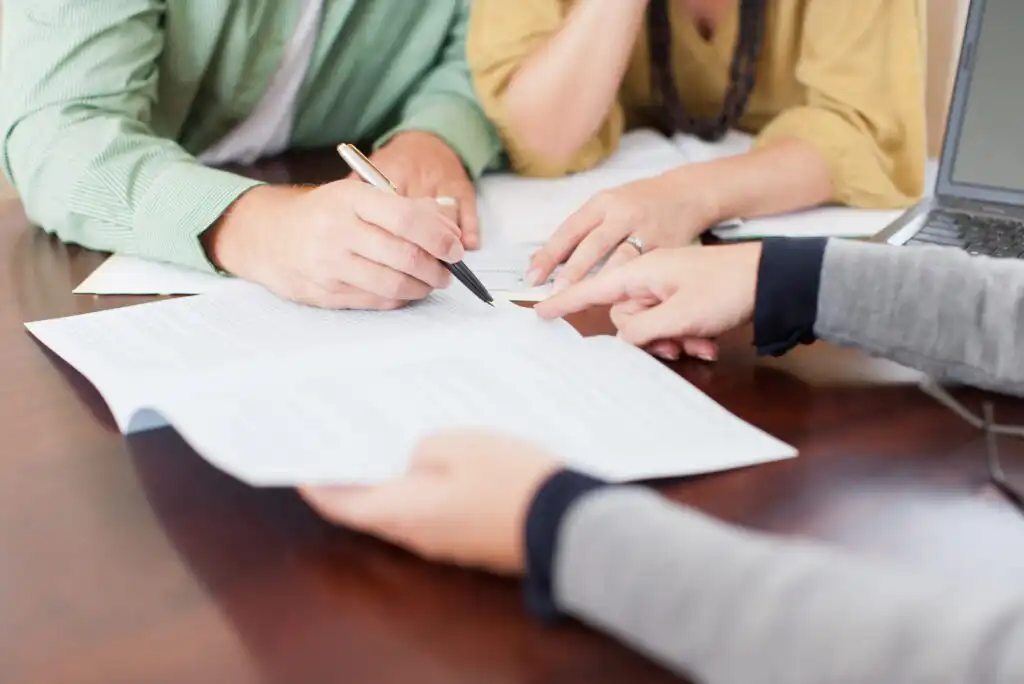 Three people sit at a table reviewing and discussing documents. One person points at the paperwork while another writes with a pen. A laptop is partially visible on the side.