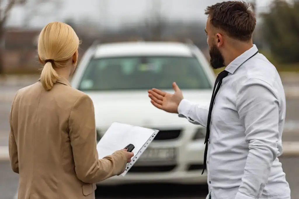 A woman holding a clipboard and car keys stands next to a man in a white shirt who gestures toward a white car parked in front of them, suggesting a discussion about the vehicle.