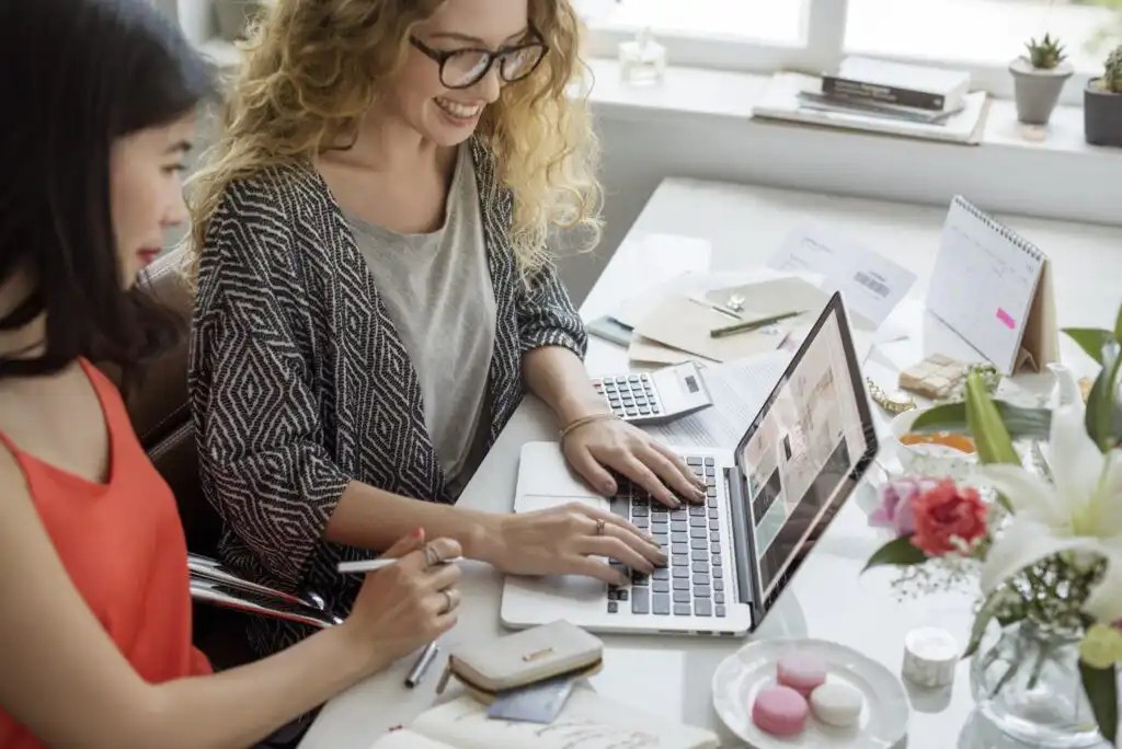 Two women sit at a bright desk, smiling while working on a laptop. The desk is arranged with notebooks, flowers, a calendar, macarons, and office supplies. Sunlight streams in through a window behind them.
