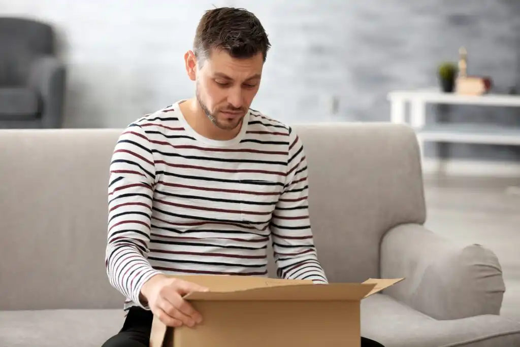 A man in a striped shirt sits on a couch, looking down with a thoughtful expression as he opens a cardboard box in a bright living room.