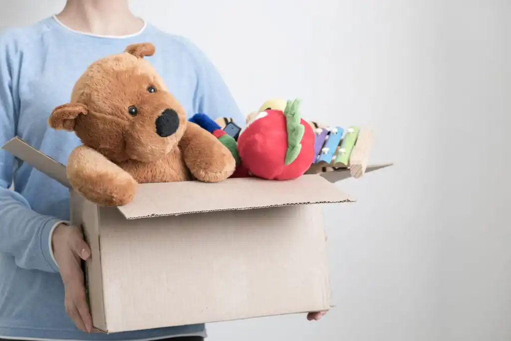 A person in a blue sweater holds an open cardboard box filled with toys, including a brown teddy bear, a plush strawberry, and colorful wooden blocks, against a plain light background.