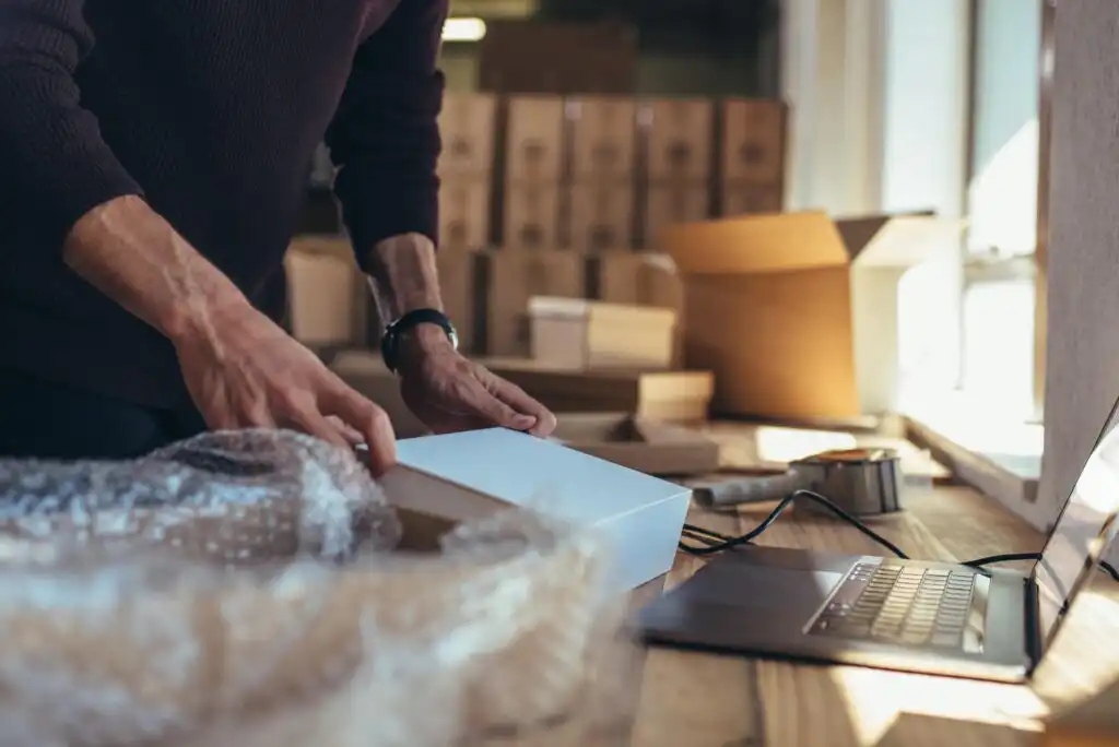 A person packing a white box on a wooden table next to a laptop, surrounded by bubble wrap and cardboard boxes in a warehouse or shipping area.