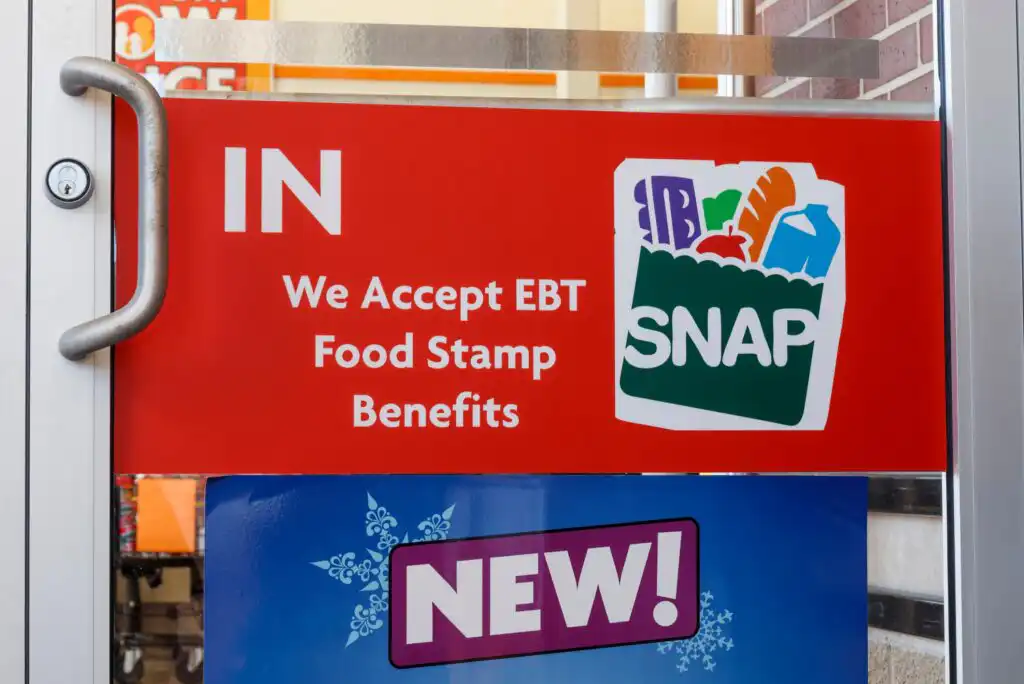 A red sign on a glass door reads IN We Accept EBT Food Stamp Benefits next to the SNAP logo, which features groceries in a bag. Below, a purple NEW! sign is partially visible.