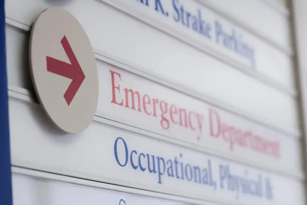 A directional sign with a red arrow points to the Emergency Department; blue text below shows directions to Occupational, Physical, and other departments. The sign appears in a medical facility.