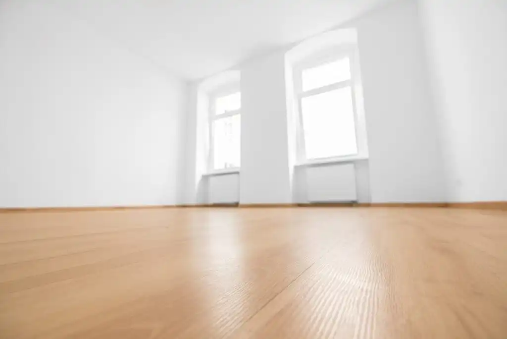 Bright, empty room with white walls, two large windows, and light wooden flooring seen from a low angle. Natural light fills the minimalist, unfurnished space.
