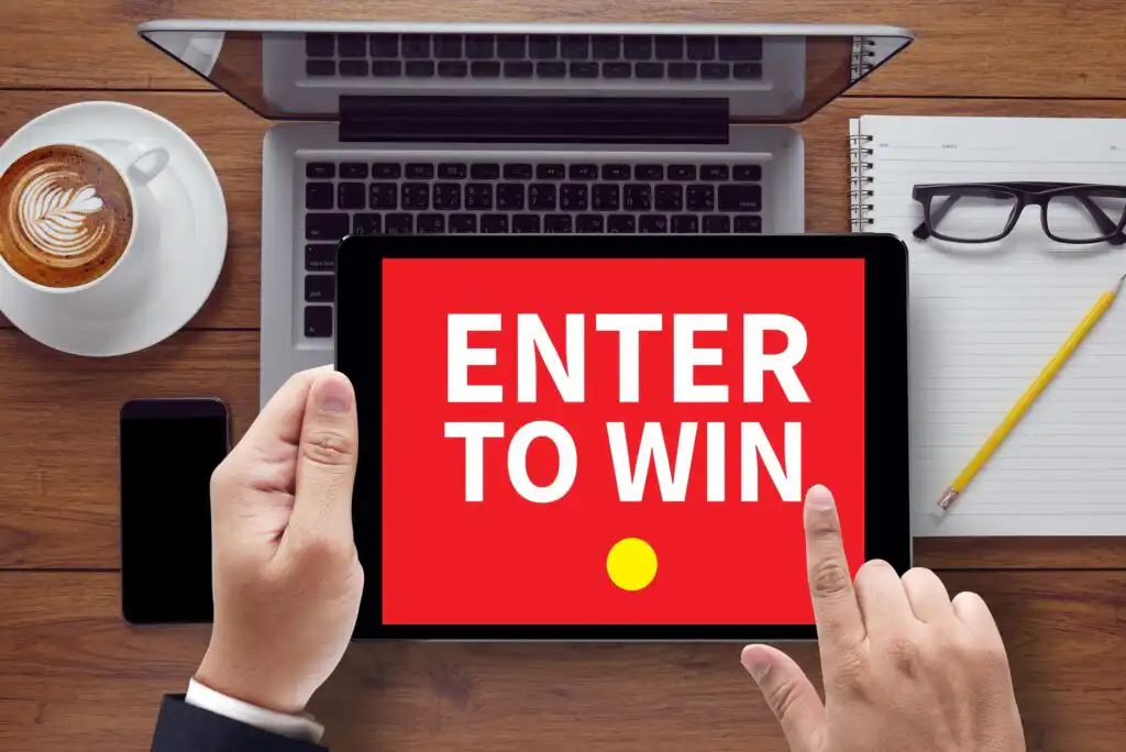 A person holds a tablet displaying a red screen with the text ENTER TO WIN. The tablet is on a desk with a laptop, coffee cup, smartphone, glasses, and a notepad with a pencil.