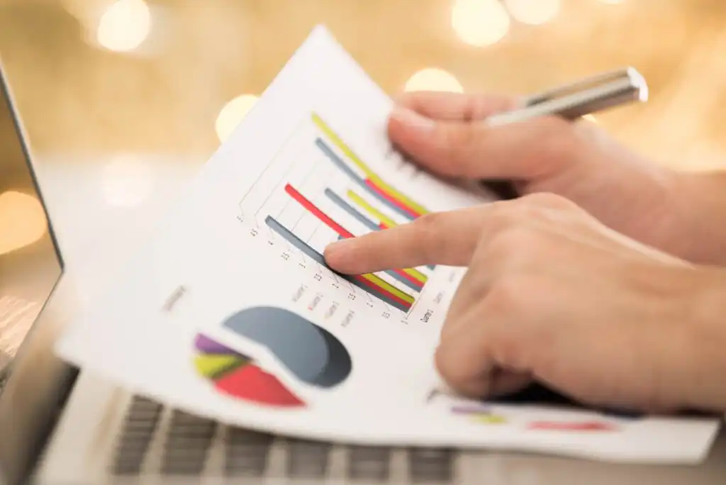 A person points at a colorful bar graph on a printed sheet while holding a pen, with a pie chart visible below. The sheet is placed on a laptop keyboard, suggesting data analysis or business work.