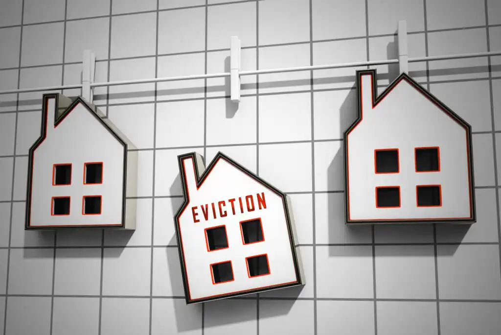 Three house-shaped objects hang on hooks against a tiled wall; the middle house is tilted and labeled EVICTION in red letters, symbolizing the issue of eviction among homes.