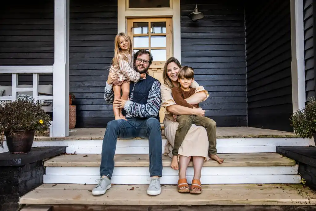 A smiling family of four sits on the steps of a dark wooden house. The father holds a young daughter, while the mother hugs a young son. They all look happy and relaxed on the porch.