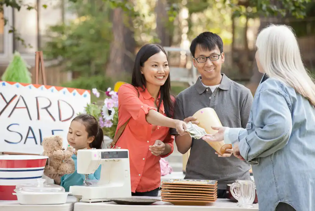 A family at a yard sale stands behind a table while an elderly woman hands money to a smiling woman. A child sits nearby playing with a teddy bear. A YARD SALE sign is visible in the background.
