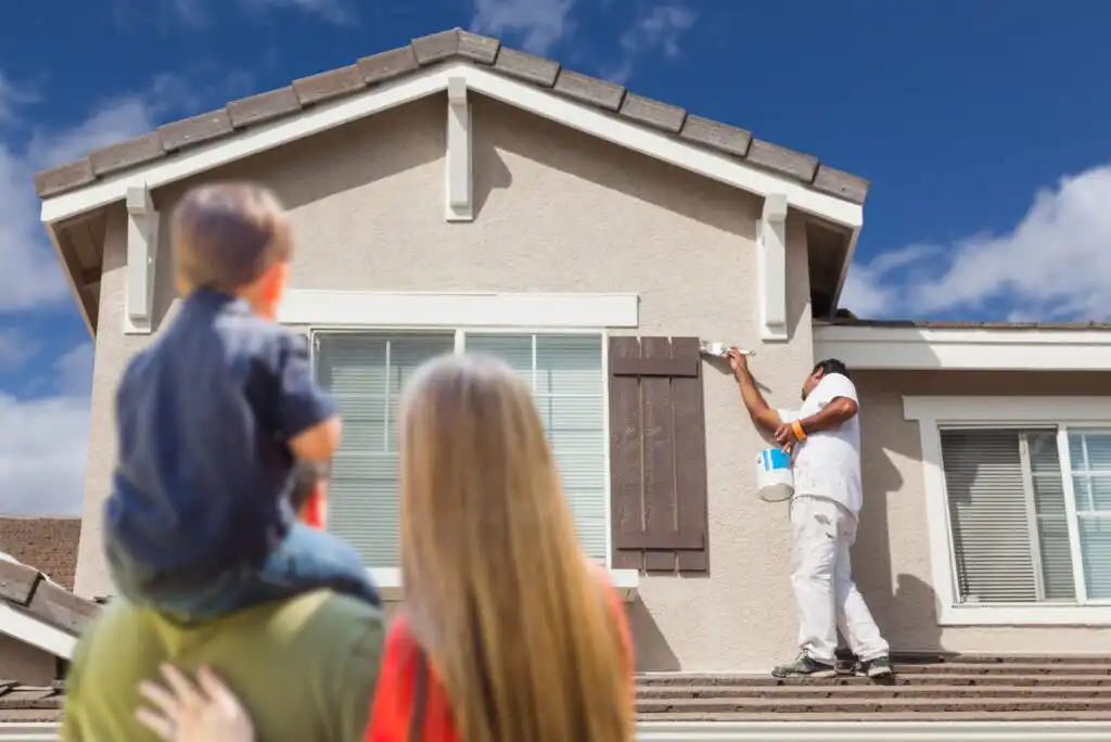 A painter applies paint to the shutters of a beige house while a woman and a child, with their backs to the camera, watch from the foreground. The sky is blue with scattered clouds.