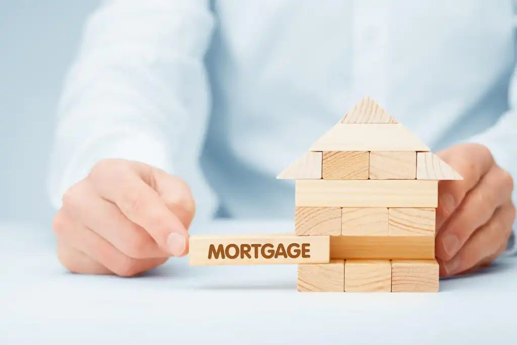 A person pulls a wooden block labeled MORTGAGE from a model house made of stacked wooden blocks, symbolizing the impact of mortgages on home stability and ownership.