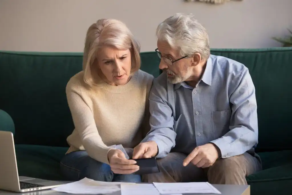An older couple sits on a couch looking concerned while reviewing bills and documents. The woman holds a calculator, and the man points at a paper on the table, with a laptop open nearby.