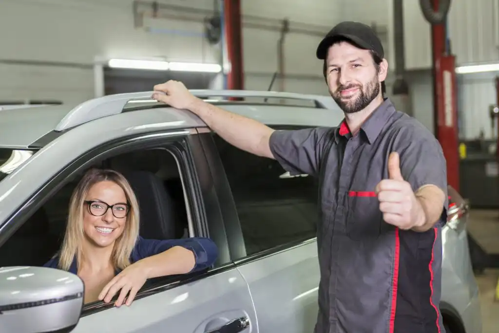 A smiling woman sits in the driver’s seat of a silver car while a mechanic in a uniform and cap stands beside her, giving a thumbs-up gesture in an auto repair shop.