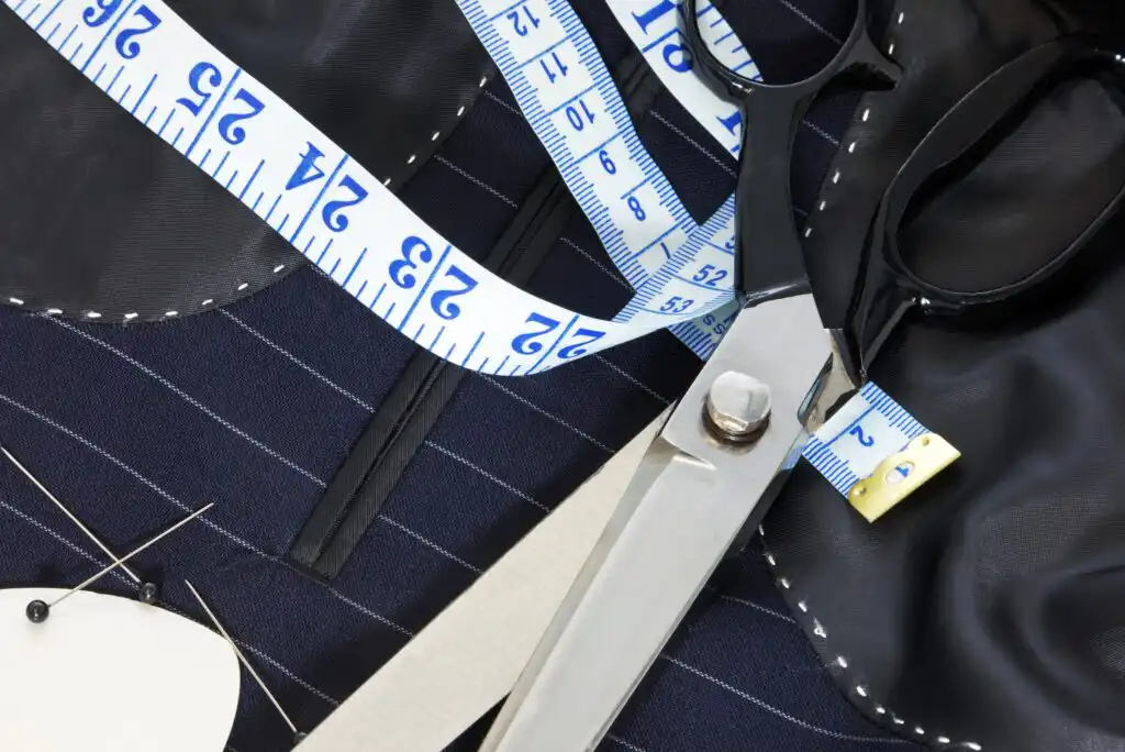 A close-up of tailoring tools, including metal scissors, a measuring tape, fabric with white pinstripes, chalk, and sewing pins, arranged on a work surface.
