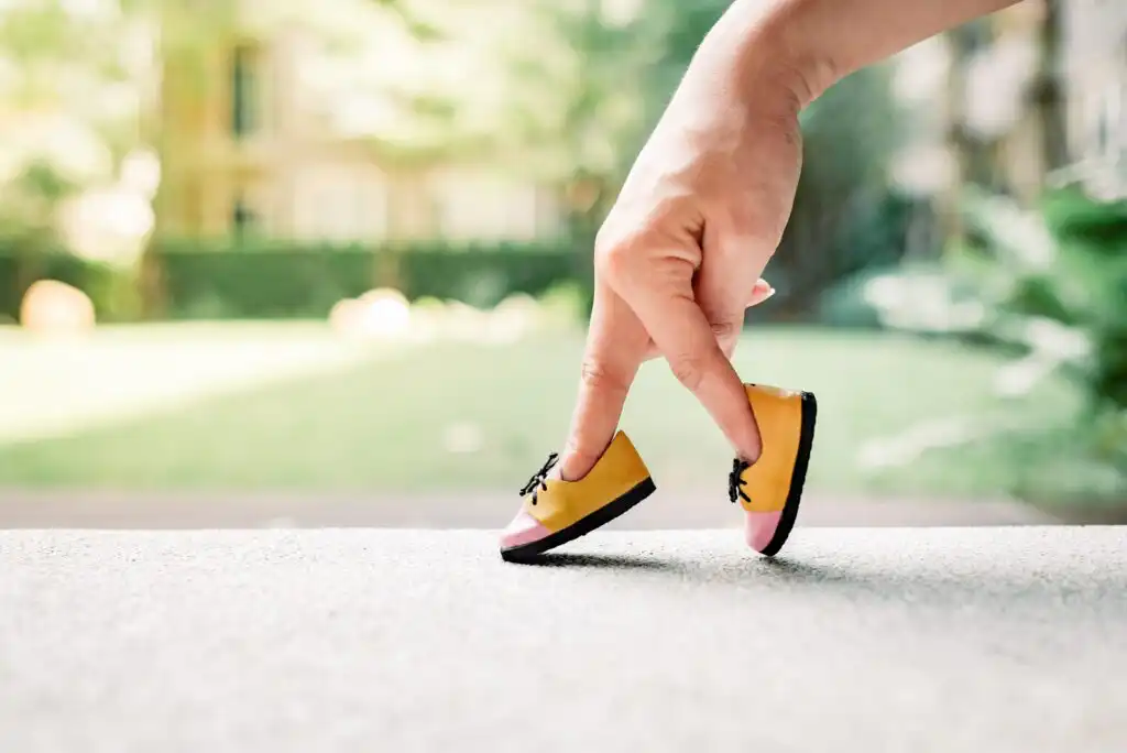 A hand with two fingers walking on the ground, wearing tiny yellow and pink shoes, mimicking legs. The background is blurred greenery outdoors.