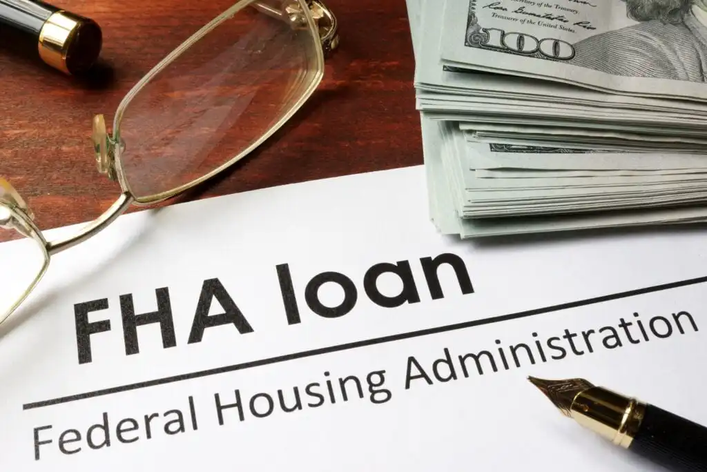 A document labeled FHA loan Federal Housing Administration lies on a desk next to a fountain pen, a pair of eyeglasses, and a stack of hundred-dollar bills.