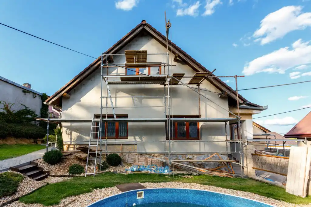 A house with scaffolding set up in front for renovation work; wooden planks and ladders are attached, and a swimming pool is visible in the foreground under a partly cloudy blue sky.