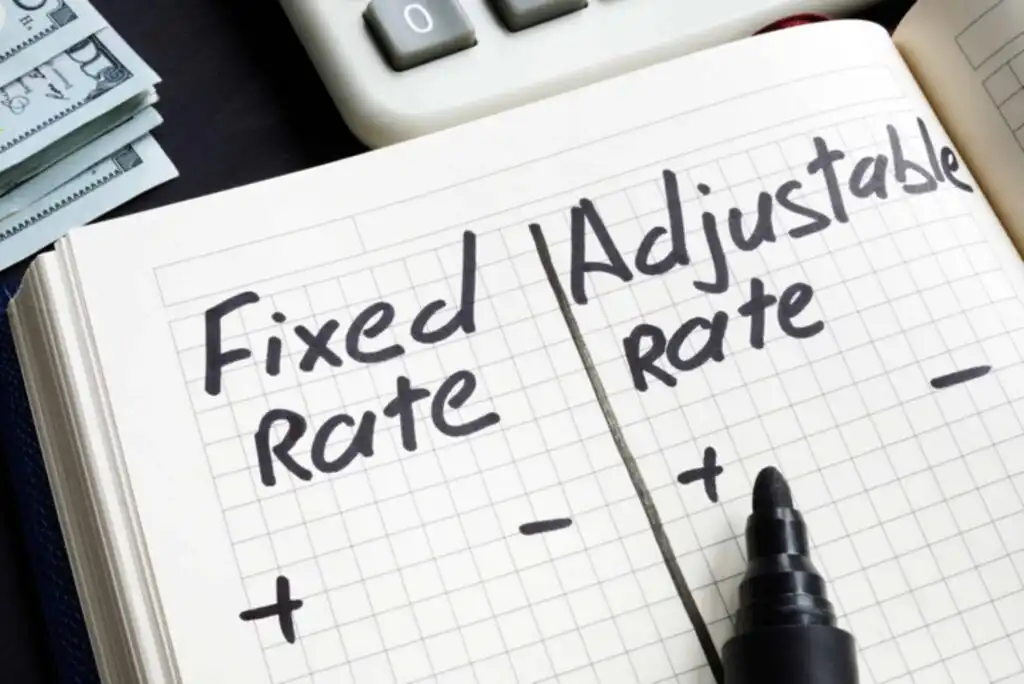A notebook shows “Fixed Rate” and “Adjustable Rate,” each with plus and minus signs underneath. A black marker rests on the page, and part of a calculator and a stack of cash are visible in the background.