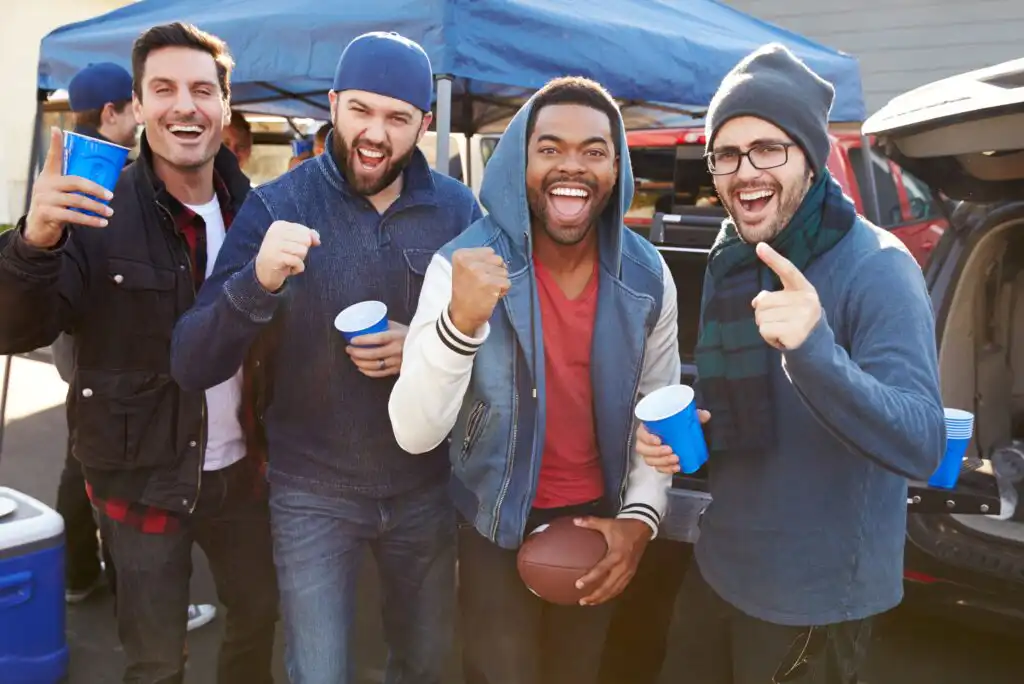 Four excited men stand together at a tailgate, smiling and cheering. Three hold blue cups, one holds a football. A blue canopy and car trunk are visible in the background. The men are dressed casually for cool weather.