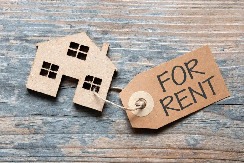 A cardboard cutout of a house is attached to a brown tag with the words “FOR RENT” written on it, placed on a rustic wooden surface.