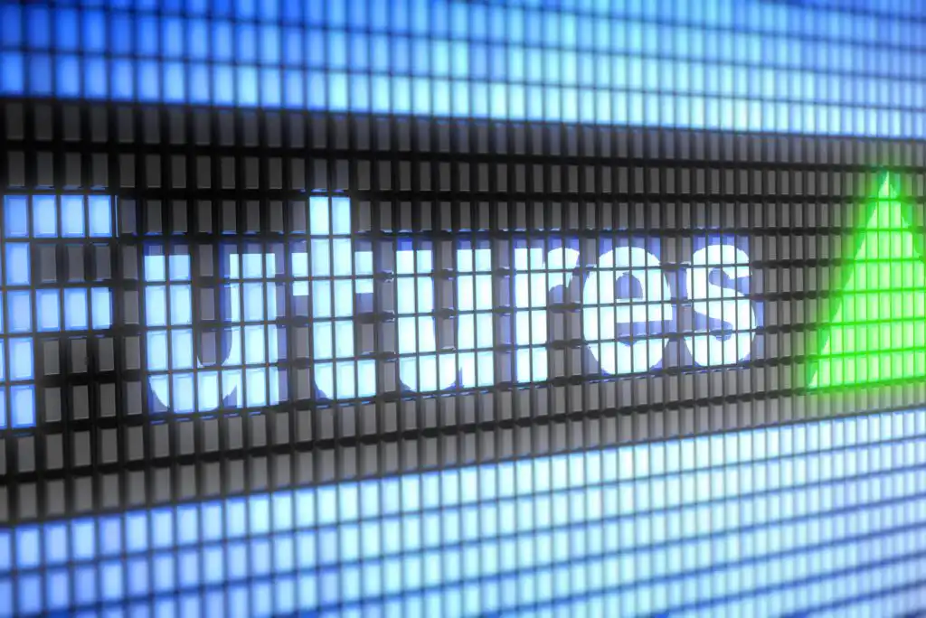 The word Futures is displayed in bright white letters on an electronic screen, next to a green upward-pointing triangle, suggesting a rise in futures markets. The background consists of illuminated digital grid pixels.